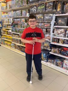 finn-stood-smiling-in-toy-shop-wearing-red-tshirt-and-blue-pants-and-holding-a-white-cane-with-a-ball-tip-225×300