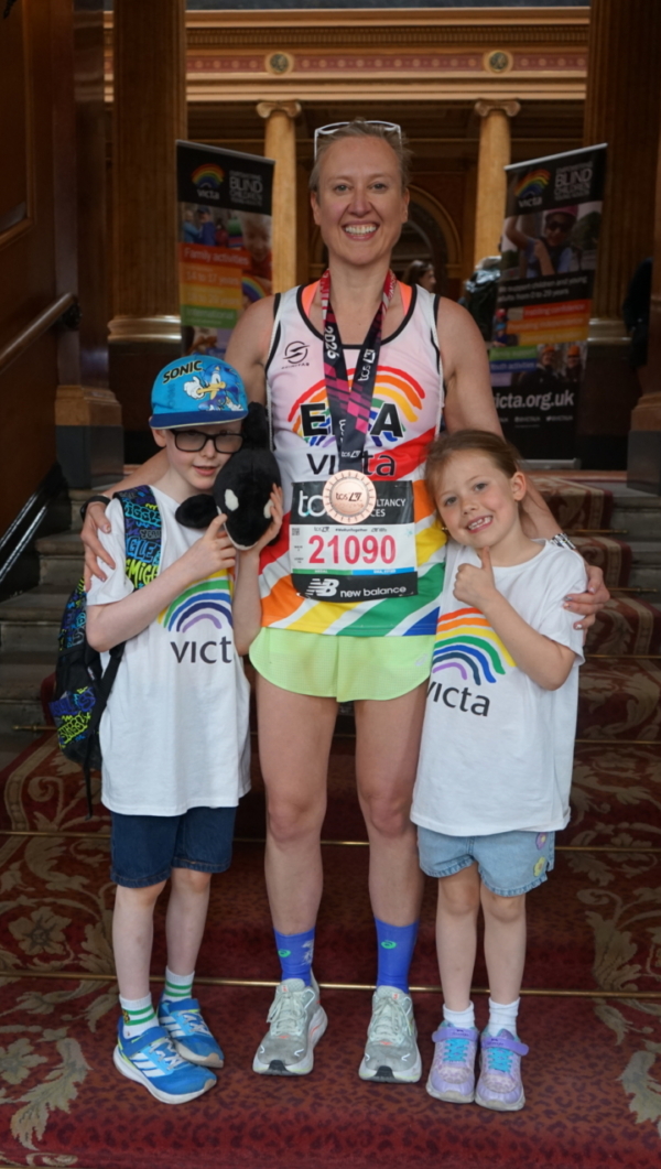 ema-party Runner stands with arms round two children at the entrance to a grand building. SHe wears a medal and VICTA running vest.
